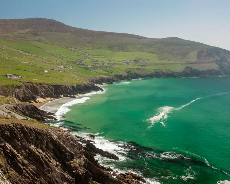 Scenic view of lush green cliffs meeting a turquoise sea, with scattered houses and a winding road along the coastline.