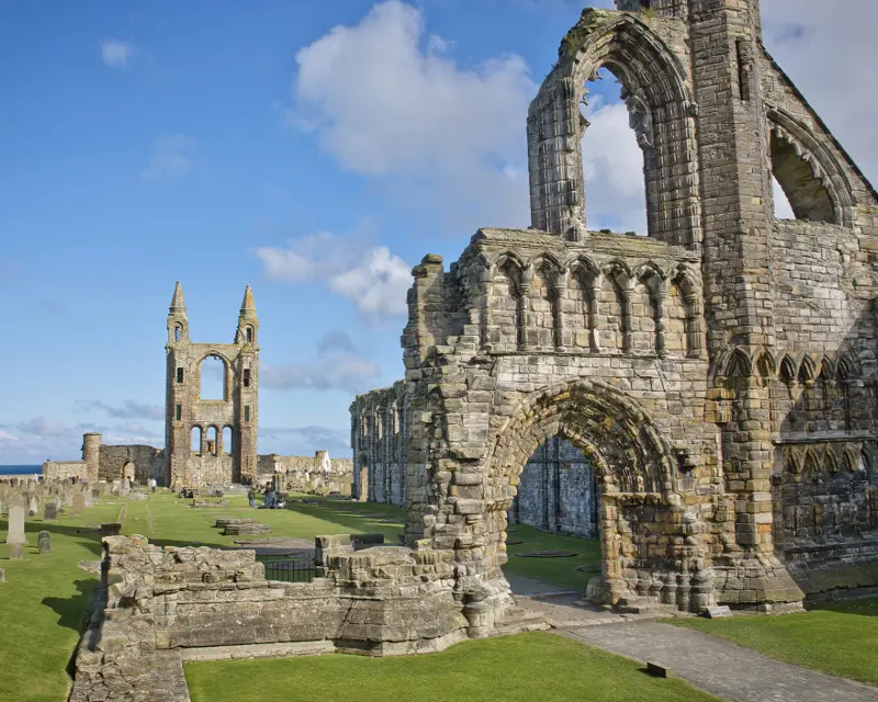 Ruins of an ancient stone abbey with towers and gravestones, set against a clear blue sky by the sea.
