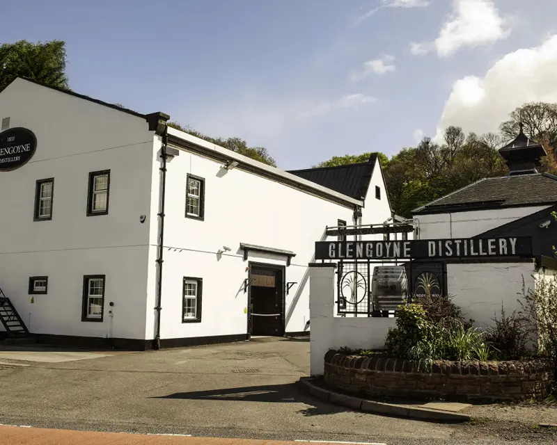 Glengoyne Distillery building with sign, lush greenery, and blue sky, inviting visitors to explore its whisky heritage.