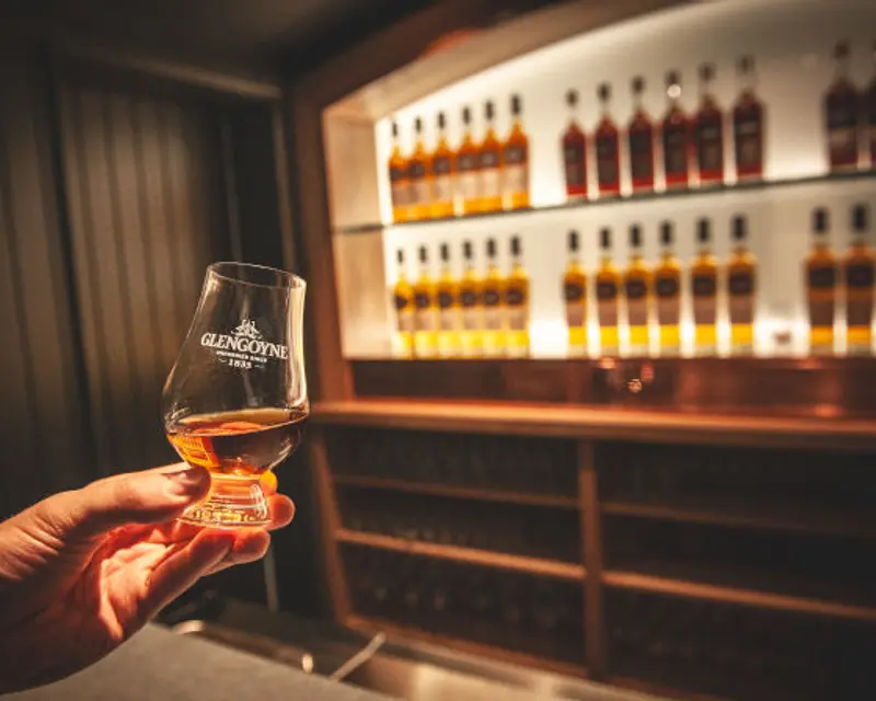 A hand holding a Glengoyne whisky glass in front of a stylish shelf filled with whisky bottles.