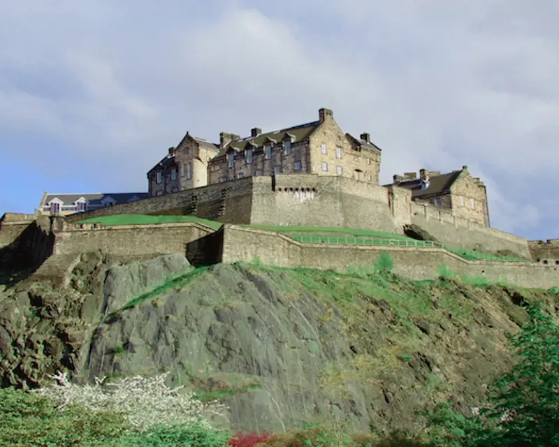 Edinburgh Castle perched atop a rocky hill, surrounded by lush greenery and dramatic skies. A historic landmark in the UK.