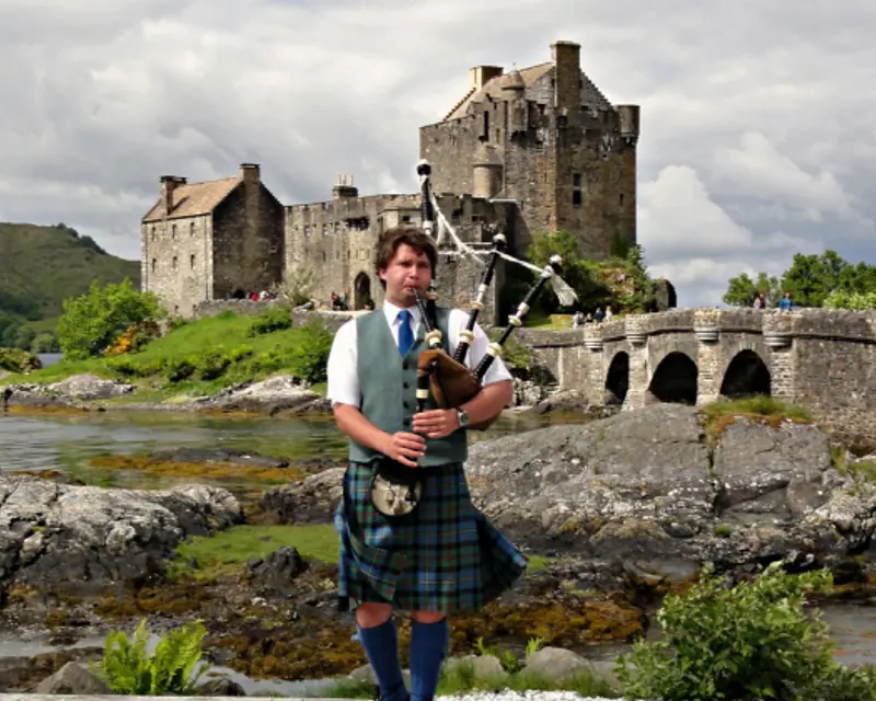 A man in a kilt plays bagpipes near a historic castle by the water, surrounded by lush greenery and dramatic skies.