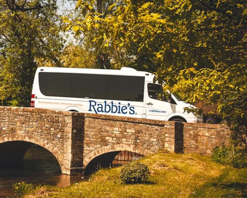 A Rabbie's Tours minibus crosses a stone bridge surrounded by lush greenery, highlighting small group travel in the UK.