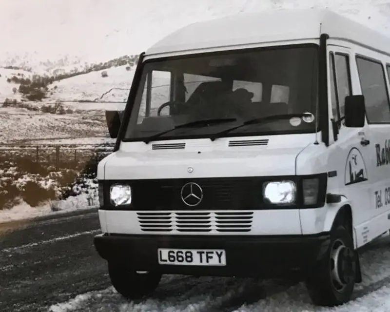 A white Mercedes van branded with Rabbie's Tours driving on a snowy road with hills in the background.