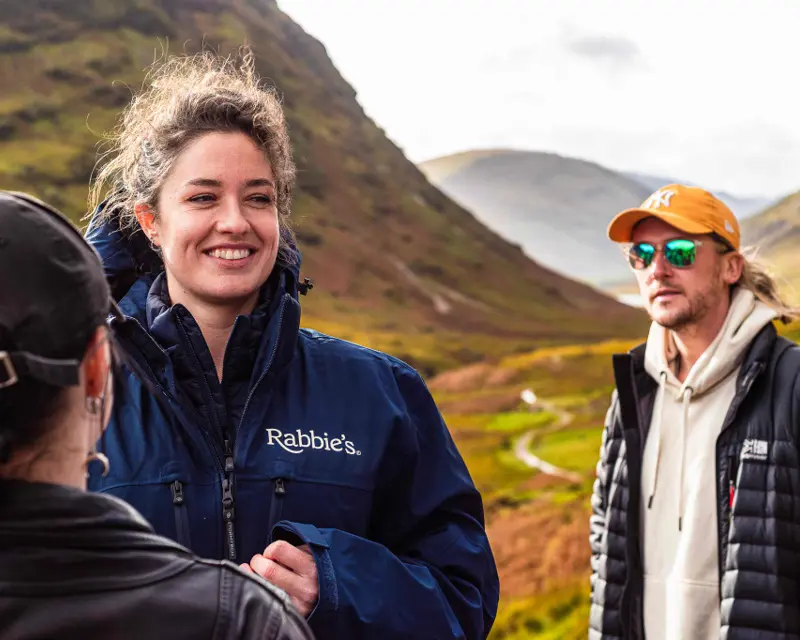 A Rabbie’s Tours guide smiles while talking to guests in a scenic UK landscape, with mountains and greenery in the background.