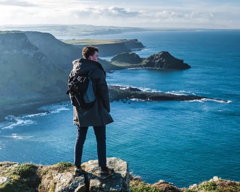 A traveler stands on a rocky ledge, overlooking stunning coastal cliffs and a vibrant blue sea under a cloudy sky.