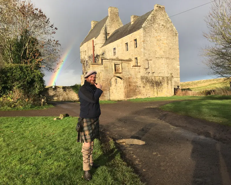 A man in a kilt poses beside a historic stone building, with a rainbow appearing in the background against a cloudy sky.