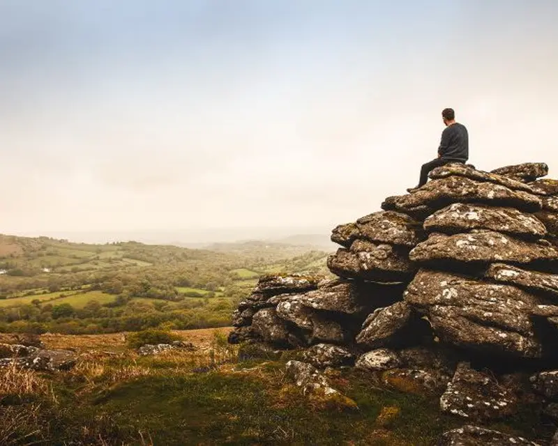 A person sits atop a stone pile, gazing over serene rolling hills and a misty landscape, embodying the spirit of exploration.
