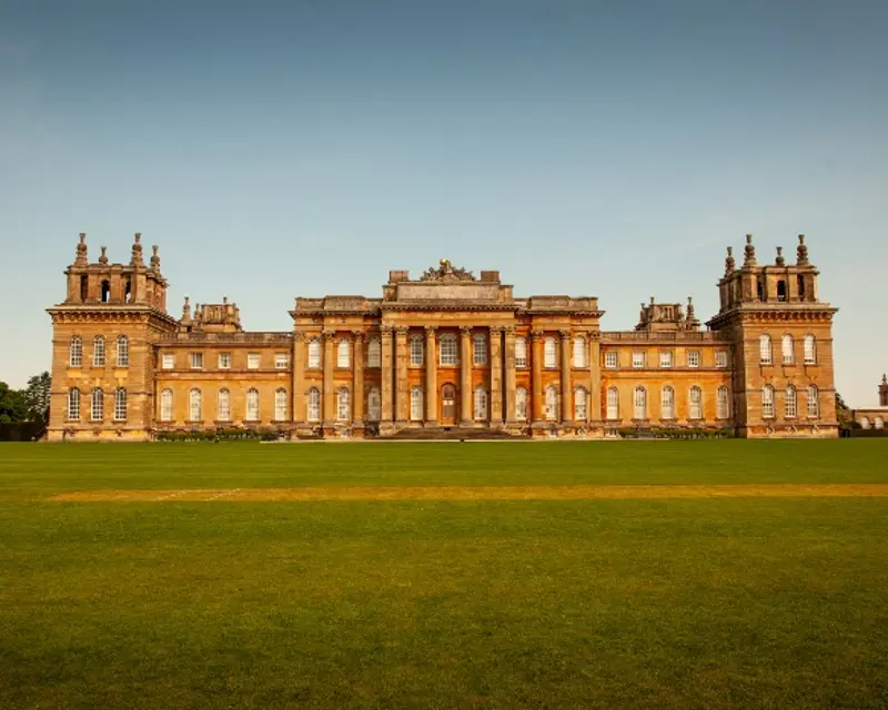 A grand historic building with intricate architecture, surrounded by lush green lawns under a clear blue sky.