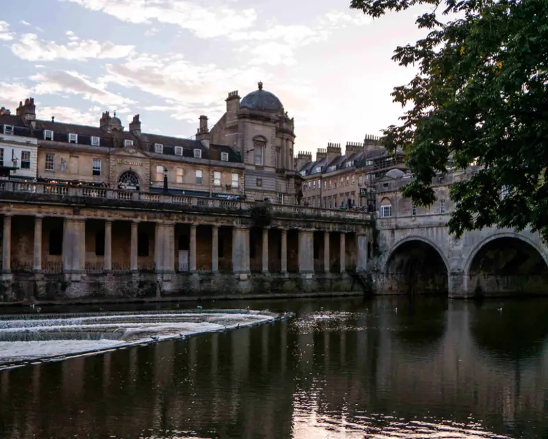 Historic buildings line the River Avon in Bath, with a stone bridge arching over the water under a serene sky.