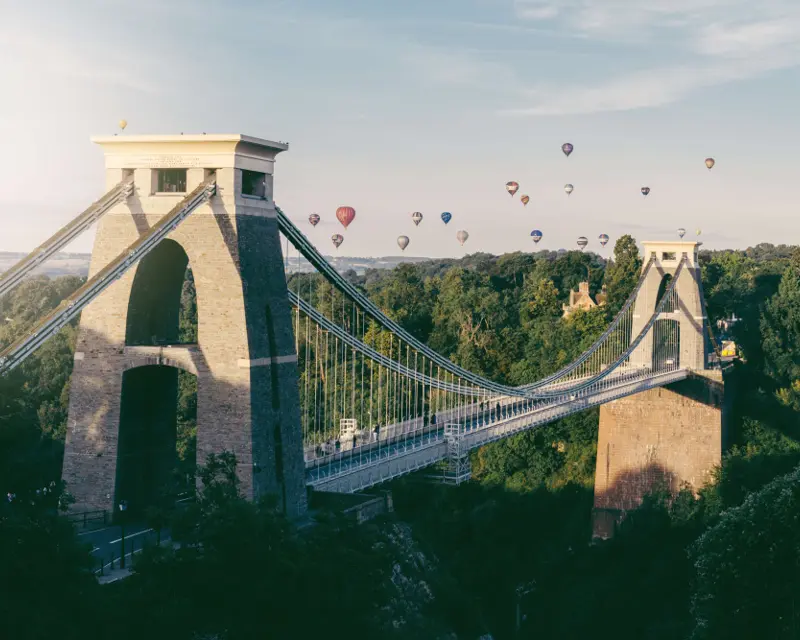 Bristol's iconic suspension bridge with hot air balloons floating above, surrounded by lush greenery in a scenic view.