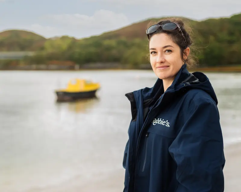A woman in a Rabbie's Tours jacket stands by the water, with a yellow boat in the background and green hills behind her.