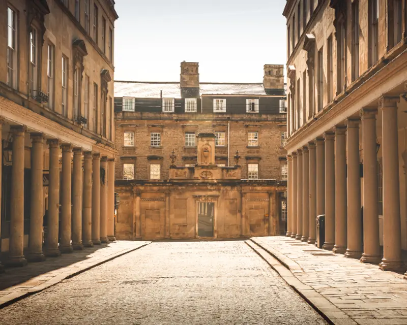 A cobblestone street flanked by historic buildings and columns, leading to an impressive facade under a clear sky.