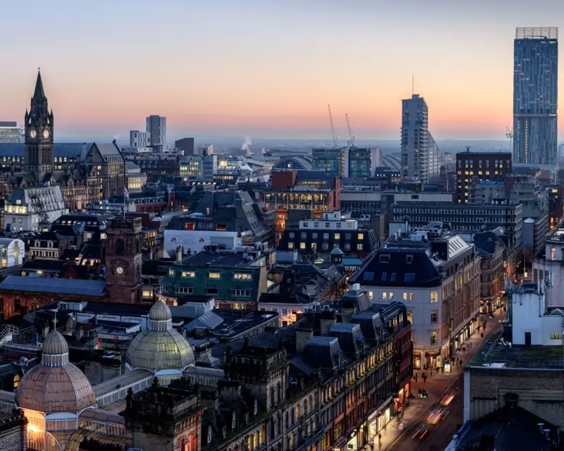 Cityscape of Manchester at dusk, showcasing historic architecture and modern buildings against a colorful sky.