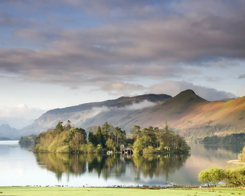 Scenic view of a tranquil lake surrounded by lush greenery and mountainous terrain under a cloudy sky.