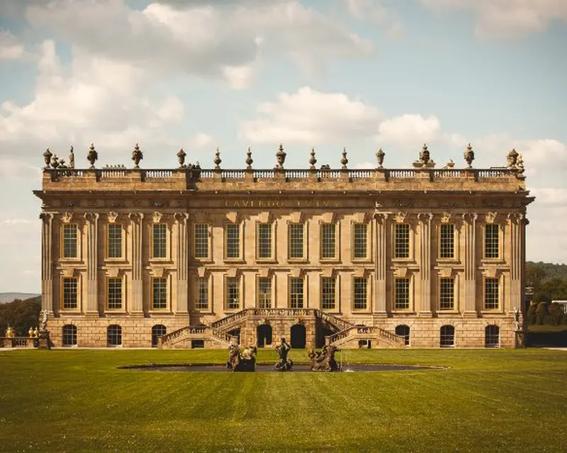 A grand, historic mansion with intricate architecture set against a clear sky, surrounded by lush green lawns.