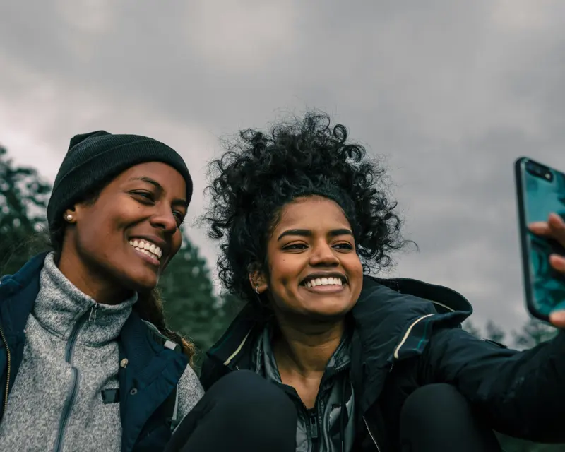 Two friends smiling and taking a selfie together outdoors, with a cloudy sky in the background.