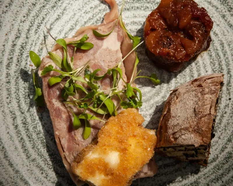 An elegant dish featuring sliced meat, garnished with microgreens, accompanied by a fruit preserve and a slice of cake.