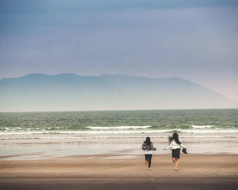 Two women run along a sandy beach, with gentle waves and misty mountains in the background, capturing a moment of adventure.