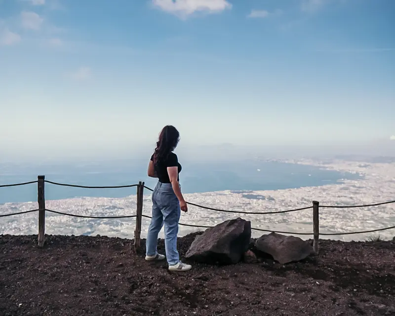 A person stands on a rocky overlook, gazing at a stunning landscape below, with distant city views and a blue sky.