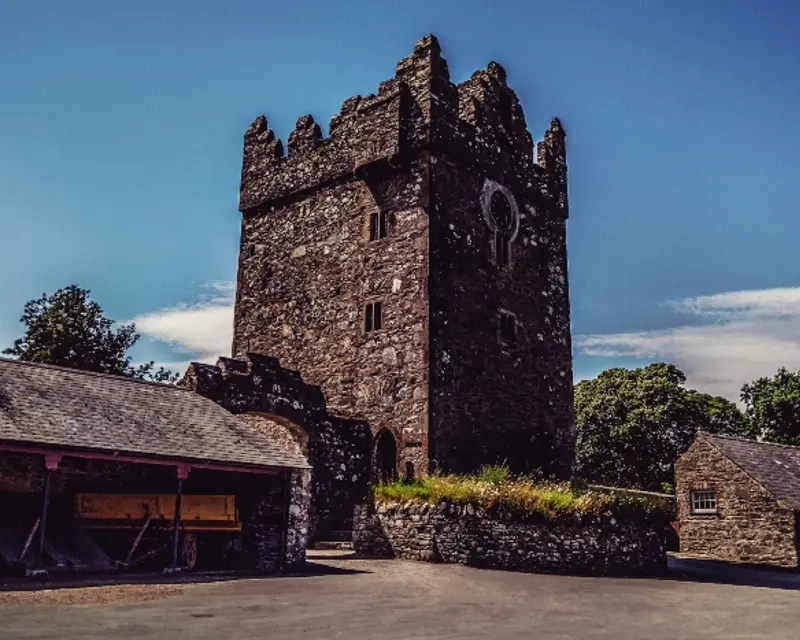 A historic stone tower with tall crenelations, surrounded by lush greenery and rustic buildings under a clear blue sky.