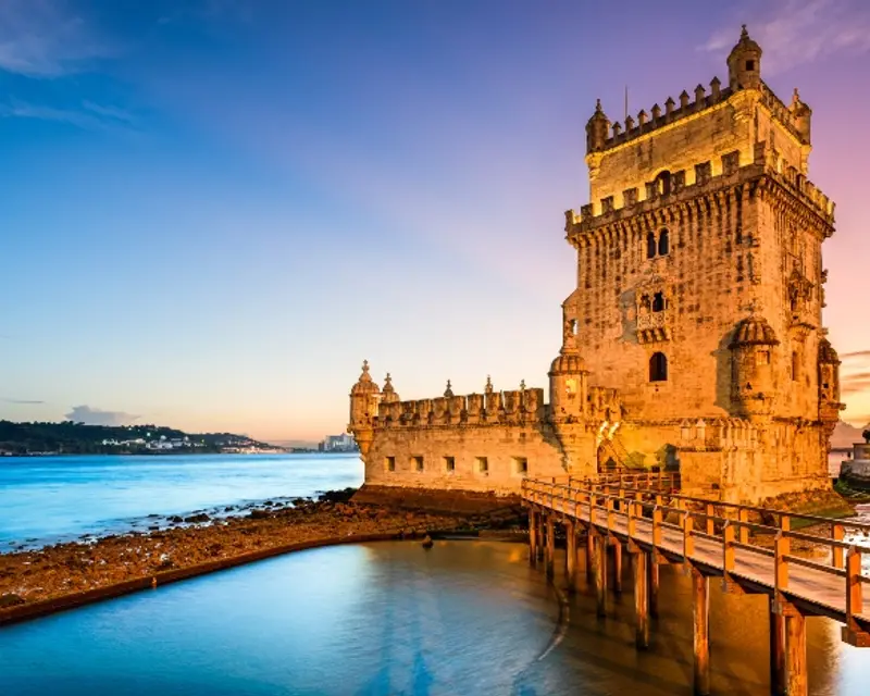Historic stone tower by the water at sunset, with a wooden walkway leading to it and a serene skyline in the background.