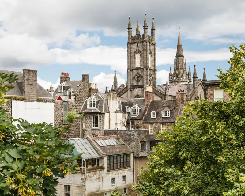 View of a historic city skyline with spires and rooftops framed by lush greenery under a cloudy sky.