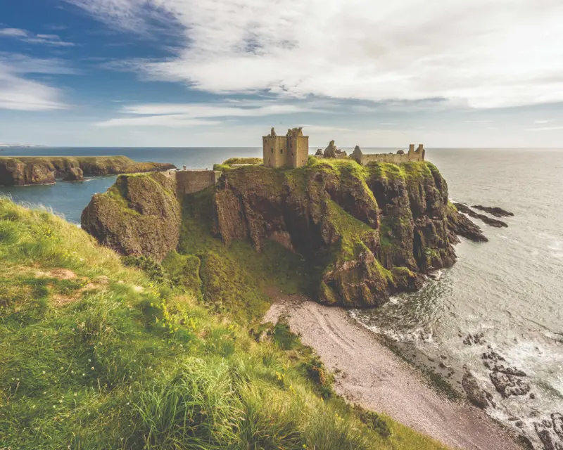 Scenic view of a coastal castle on a cliff, surrounded by lush greenery and the ocean under a cloudy sky.