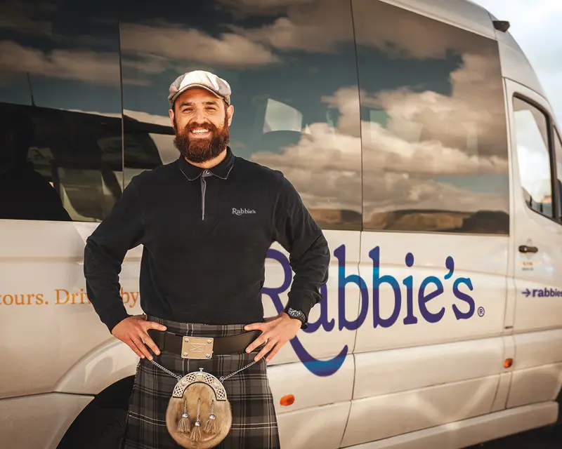 A smiling tour guide in a kilt stands in front of a Rabbie's Tours van, promoting small group tours in the UK.
