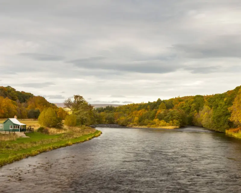 Scenic view of a river winding through colorful autumn trees, with a quaint green house along the riverbank.
