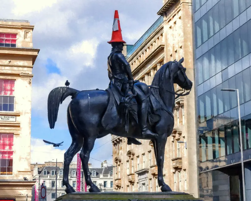 A statue of a horseman wearing a traffic cone on his head, set against urban buildings and a blue sky.