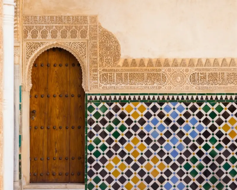 Intricate carved archway with a wooden door beside a colorful tiled wall, showcasing stunning geometric patterns.