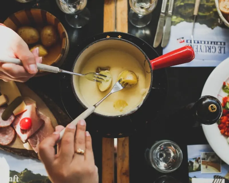 A close-up of hands dipping potatoes into cheese fondue, surrounded by various food items on a table.