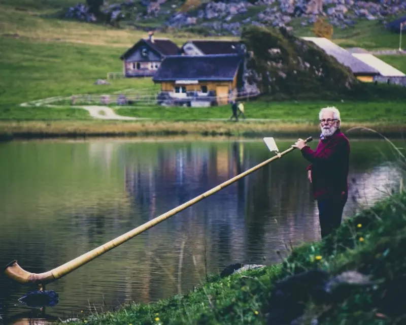 A man plays a long wooden instrument by a tranquil lake, with green hills and rustic buildings in the background.