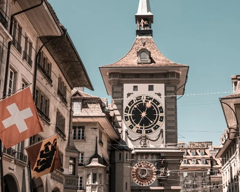 Historic clock tower in a quaint European town, flanked by flags and charming buildings under a clear blue sky.
