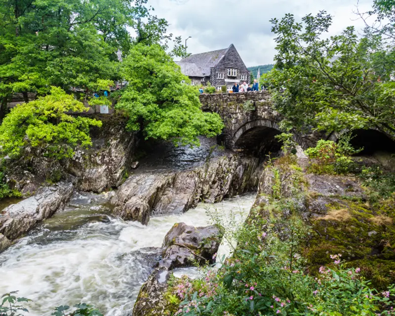 A scenic view of a rocky river with a historic bridge, surrounded by lush greenery and a charming building in the background.