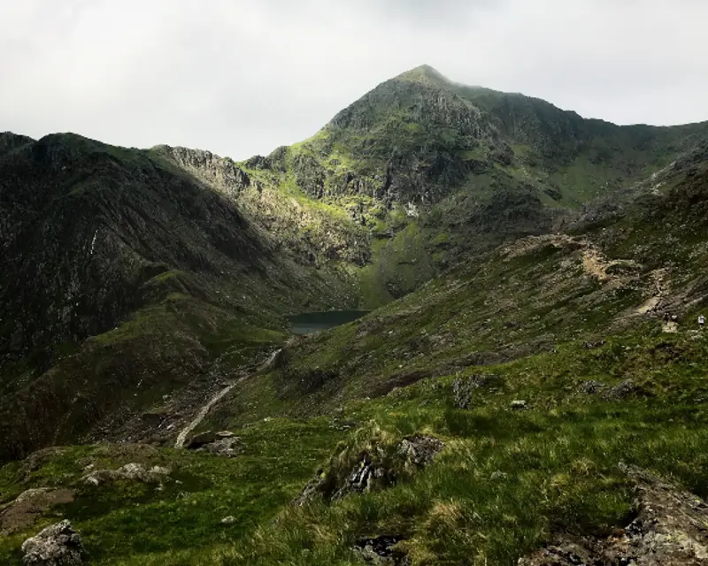 A dramatic mountain landscape with green slopes, rocky terrain, and a serene lake nestled in the valley under a cloudy sky.