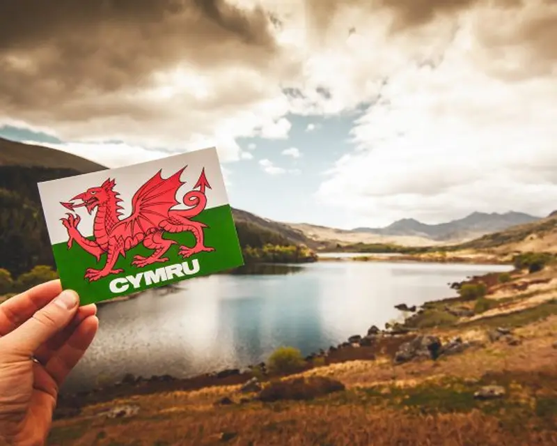 A hand holds a postcard of the Welsh flag in front of a scenic lake and mountains under a cloudy sky in Wales.