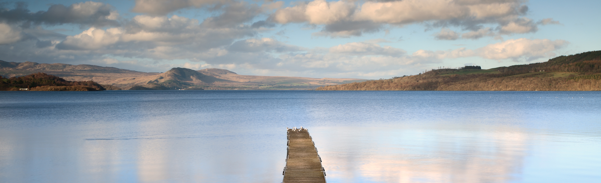 a pontoon stretching out into the still and blue waters of Loch Lomond with hills in the background