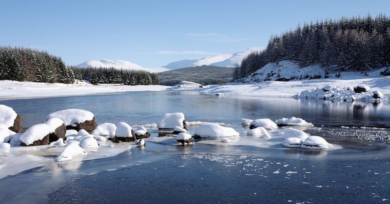 the frozen water of Loch Laggan surrounded by snow covered mountains and pine trees