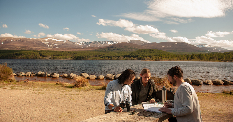 a group of people sitting at a bench in the Cairngorms national park with a loch and hills behind them