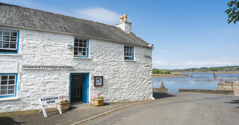 The Kirkcudbright Harbour Cottage Gallery is a picturesque white stone building with a bright blue door and window panes.