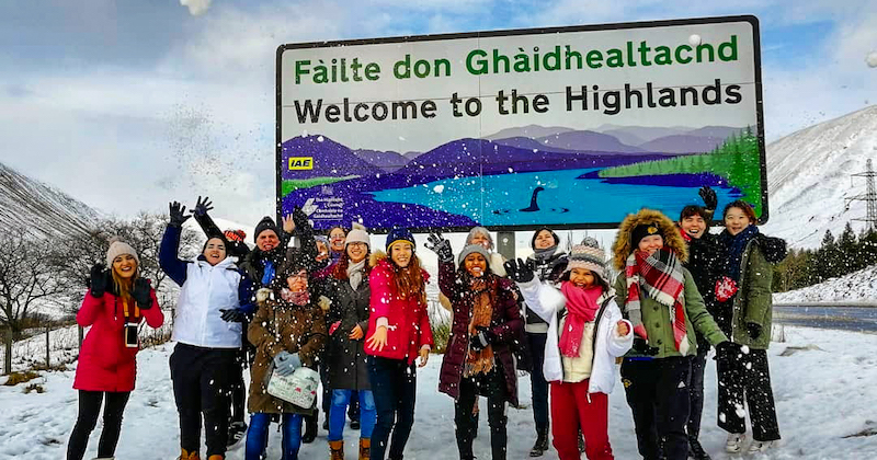 a Rabbie's tour group throwing snow in front of a 'Welcome to the Scottish Highlands' sign