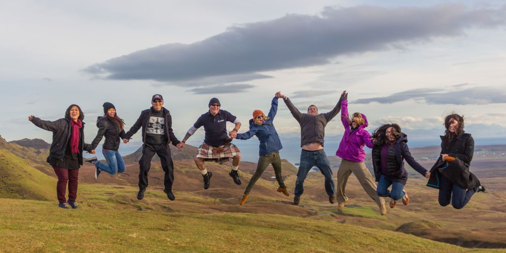 A group of peaple holding hands and jumping in air at the top of a Scottish mountain 