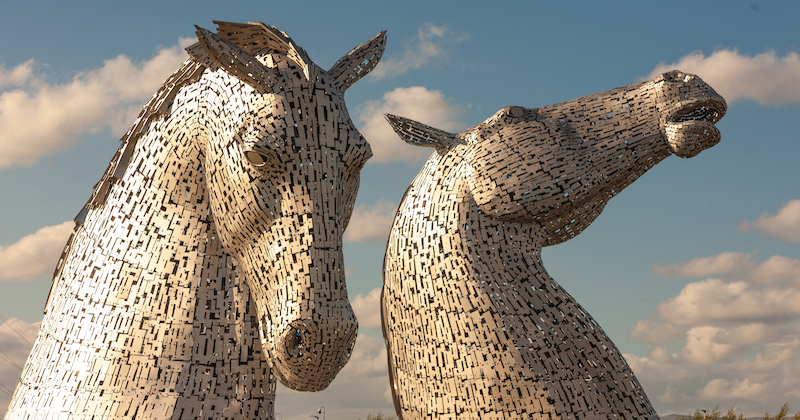 the large silver metal sculptures of the kelpies under a blue sky with fluffy white clouds