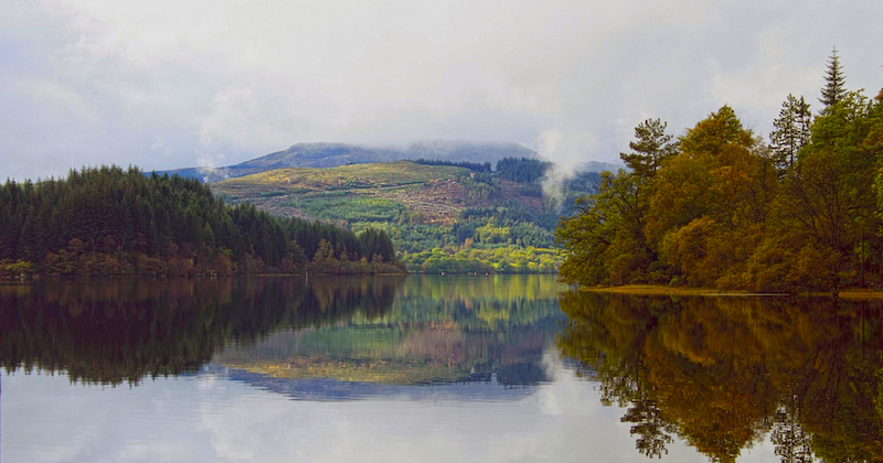 the still waters of Loch Lomond reflects the surrounding hills and forests under a misty sky