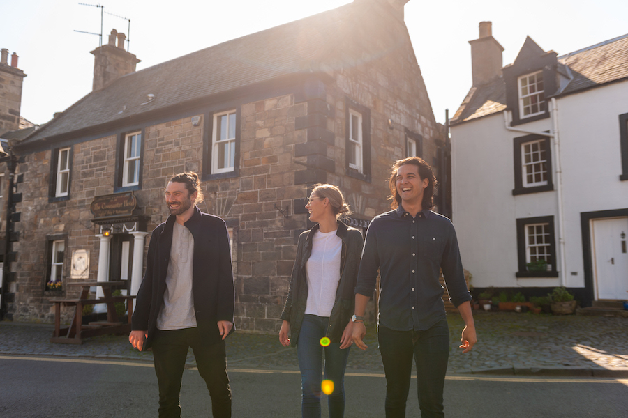 three friends outside their brown building travel accommodation in Scotland with the sun setting behind them